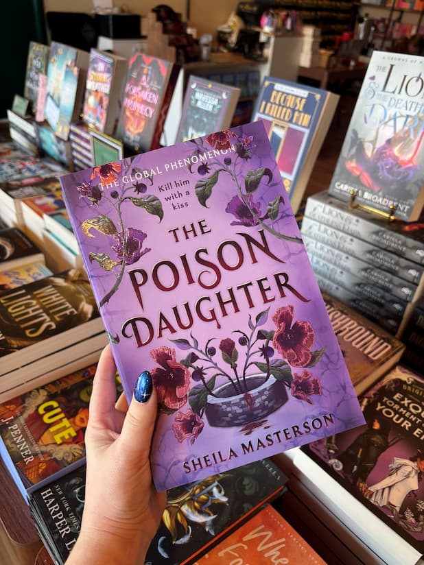 A hand with blue glitter nail polish holds the book 'The Poison Daughter' by Sheila Masterson, featuring a purple cover with floral illustrations, in a bookshop setting with other books visible in the background.