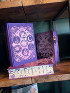 The special edition version of 'The Poison Daughter' by Sheila Masterson with the dust cover removed showcasing  purple foiling with six elemental symbols in a circle on the cover. The sprayed edges show the text "I would rather break the world than let you go". The book sits on a wooden shelf with a green background. 