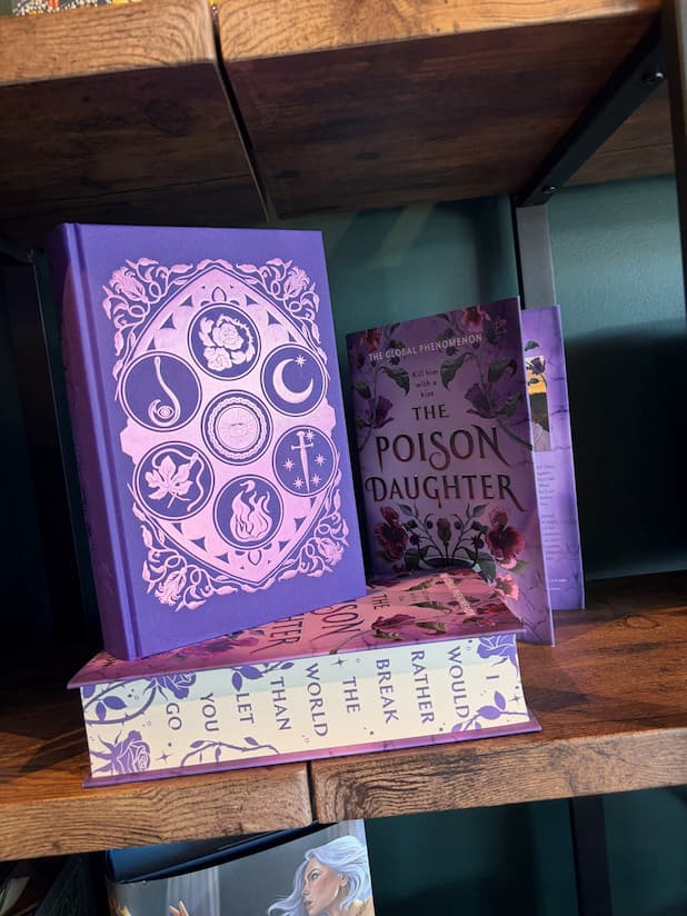 The special edition version of 'The Poison Daughter' by Sheila Masterson with the dust cover removed showcasing  purple foiling with six elemental symbols in a circle on the cover. The sprayed edges show the text "I would rather break the world than let you go". The book sits on a wooden shelf with a green background. 
