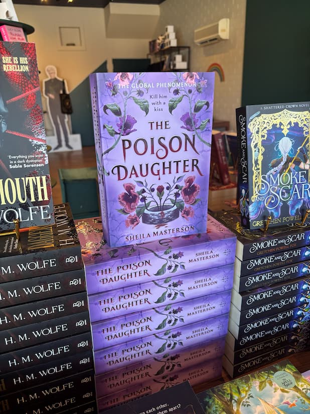 A stack of the book 'The Lion & the Deathless Dark' by Carissa Broadbent in a book store, with other book spines visible in the background.