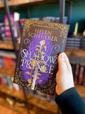 A hand holds a paperback copy of 'The Shadow Prince' by Helen Scheuerer, featuring a dark purple and black background with ornate gold filigree, a silver sword, and white title lettering. 