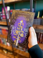 A hand holds a paperback copy of 'The Shadow Prince' by Helen Scheuerer, featuring a dark purple and black background with ornate gold filigree, a silver sword, and white title lettering. 