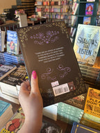 A hand with pink nail polish holds the back of a hardcover copy of 'The Shadow Prince' by Helen Scheuerer, featuring a black background with ornate gold and purple filigree with white text.