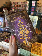 A hand with pink nail polish holds a hardcover copy of 'The Shadow Prince' by Helen Scheuerer, featuring a dark purple and black background with ornate gold filigree, a silver sword, and white title lettering. 