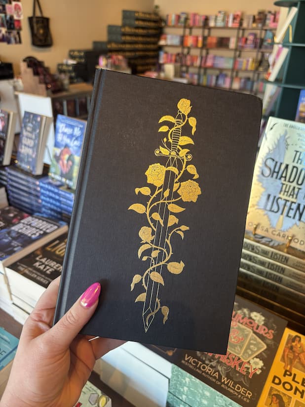A hand with pink nail polish holds a hardcover copy of 'The Shadow Prince' by Helen Scheuerer without the dust jacket, featuring a black background with a gold sword surrounded by flowers.
