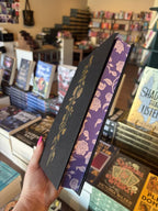 A hand with pink nail polish holds a hardcover copy of 'The Shadow Prince' by Helen Scheuerer without the dust jacket, featuring a black cover with gold accents and purple floral spray edges.