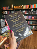A hand with pink nail polish holding the back of the book 'The Shadows That Listen' by Louisa Carmody, featuring a black background, white feathres, and white and yellow text.
