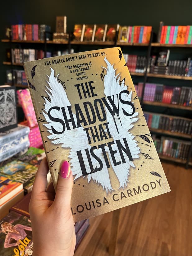 A hand with pink nail polish holds up the book 'The Shadows That Listen' by Louisa Carmody. The book cover is gold color with a large white wing design and black text.
