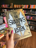 A hand with pink nail polish holds up the book 'The Shadows That Listen' by Louisa Carmody. The book cover is gold color with a large white wing design and black text.