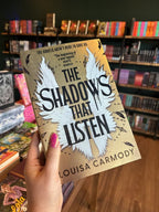 A hand with pink nail polish holds up the book 'The Shadows That Listen' by Louisa Carmody. The book cover is gold color with a large white wing design and black text.