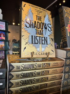 A stack of copies of the book 'The Shadows That Listen' by Louisa Carmody, with one copy standing upright on a display stand in a bookstore setting. The cover is gold with white wings and large black text in the centre.