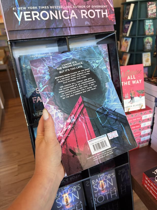 A hand with nude nail polish holding the bcak of a copy of 'The Sixth Faction' by Veronica Roth, featuring a dramatic sci-fi scene with white text in front of a swirling blue vortex.