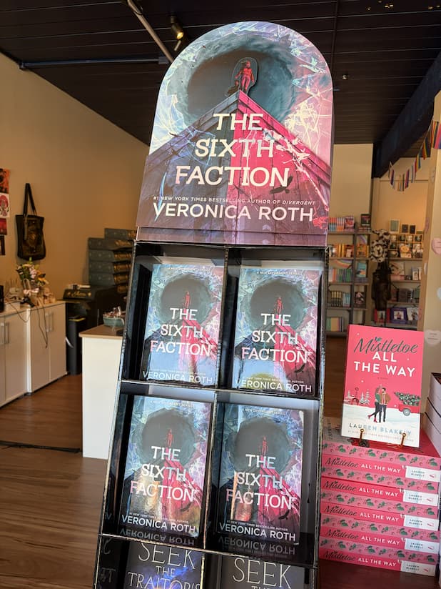 A book display stand in a bookstore showing multiple copies of 'The Sixth Faction' by Veronica Roth, with a larger version of the book cover mounted above the stand. 