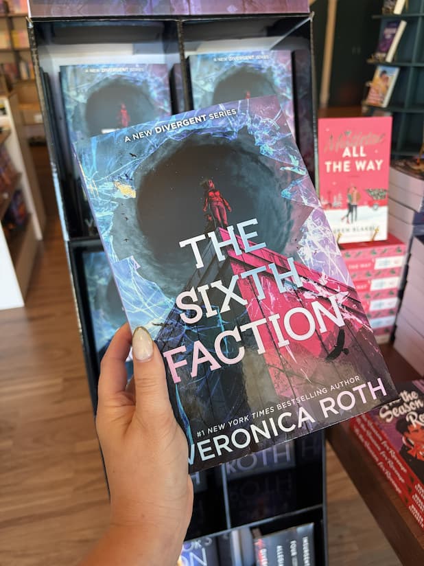 A hand with nude nail polish holding a copy of 'The Sixth Faction' by Veronica Roth, featuring a dramatic sci-fi scene with two figures standing atop a concrete building in front of a swirling blue vortex, with pink and white title text across the center.