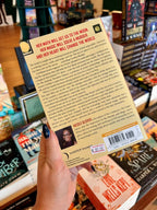 A hand with black glitter nail polish holds the back cover of a book titled 'The Starseekers' by Nicole Glover in a bookstore. The book cover is a light yellow color with black text and a barcode, with other books visible in the blurred background.