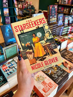 A hand holds the book 'The Starseekers' by Nicole Glover featuring a teacher in a classroom, with other book covers visible in the background.