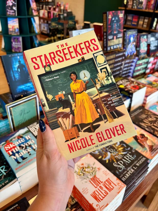 A hand holds the book 'The Starseekers' by Nicole Glover featuring a teacher in a classroom, with other book covers visible in the background.