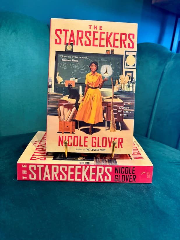 A book titled 'The Starseekers' by Nicole Glover is displayed upright on a teal couch, supported by two small gold bookends. The cover features a teacher in a classroom.