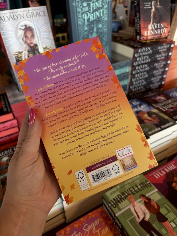 A hand with pink nail polish holding the back of a copy of 'The Sugar Maple Inn' by Laura Bloom, featuring a cozy autumnal setting.