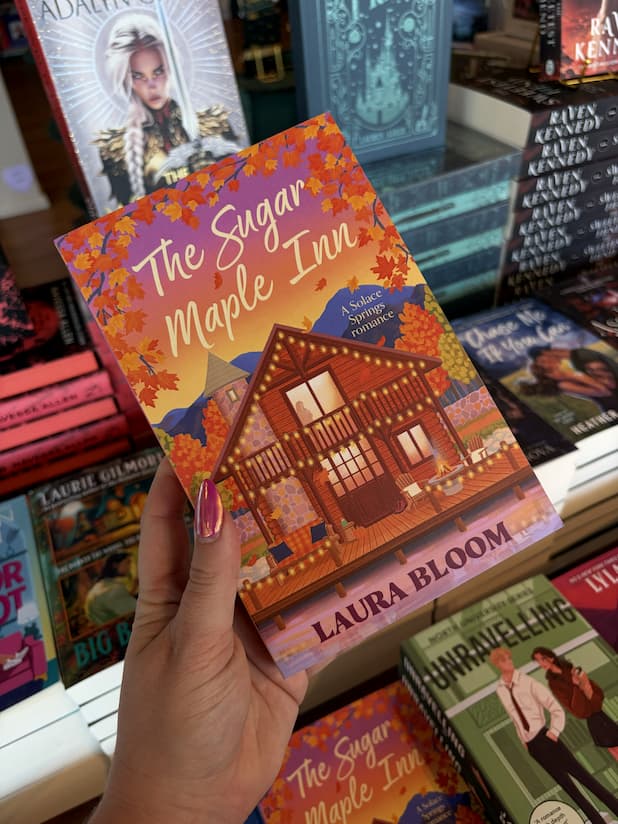 A hand with pink nail polish holding a coyp of 'The Sugar Maple Inn' by Laura Bloom, featuring a cozy cabin in an autumn setting.