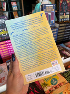 A hand with dark glittery nail polish holds the back cover of a book titled 'The Summer Girlfriend' by author Kristina Forest, with a bookstore shelf of other books in the background.