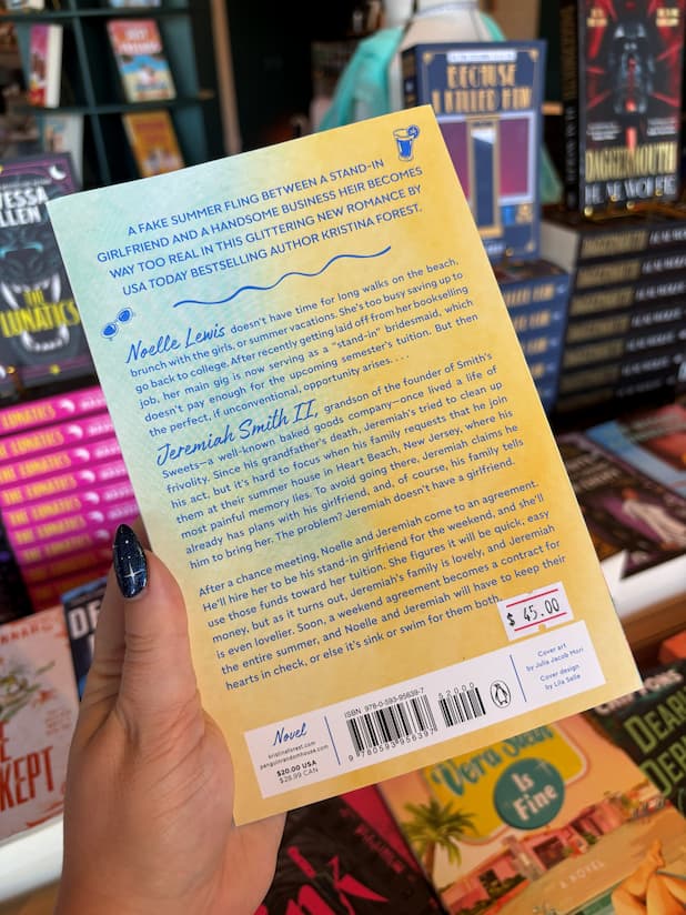 A hand with dark glittery nail polish holds the back cover of a book titled 'The Summer Girlfriend' by author Kristina Forest, with a bookstore shelf of other books in the background.