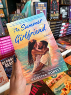 A hand holding a copy of the book 'The Summer Girlfriend' by Kristina Forest, Book One in the Heart Beach Series, the illustrated cover art features a black couple embracing on a beach with a sunset.