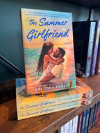 A copy of the book 'The Summer Girlfriend' by Kristina Forest displayed on a wooden bookshelf, standing upright on a small gold book stand next to other copies of the book.