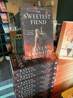 A stack of 'The Sweetest Fiend' by Raven Kennedy, featuring a woman in a white dress and dark hair against a fiery background raising her pointer finger to the brightest star in the sky.