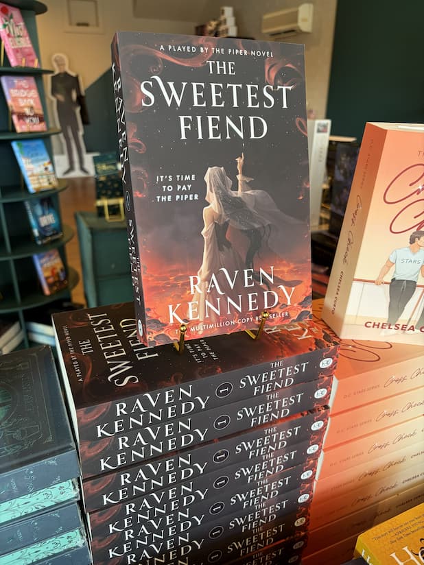 A stack of 'The Sweetest Fiend' by Raven Kennedy, featuring a woman in a white dress and dark hair against a fiery background raising her pointer finger to the brightest star in the sky.