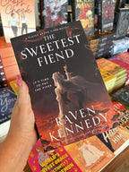 A hand with pink nail polish holds a copy of 'The Sweetest Fiend' by Raven Kennedy, featuring a woman in a white dress and dark hair against a fiery background raising her pointer finger to the brightest star in the sky.