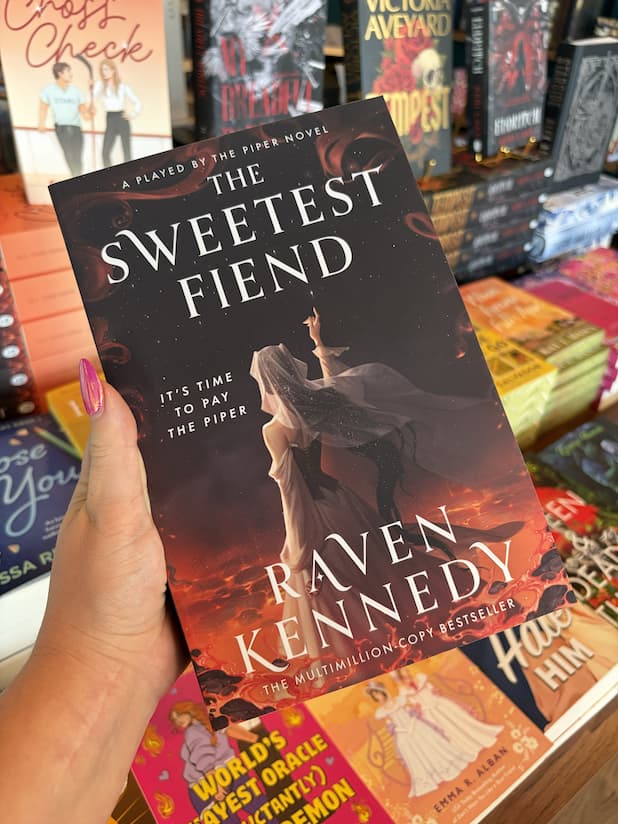 A hand with pink nail polish holds a copy of 'The Sweetest Fiend' by Raven Kennedy, featuring a woman in a white dress and dark hair against a fiery background raising her pointer finger to the brightest star in the sky.