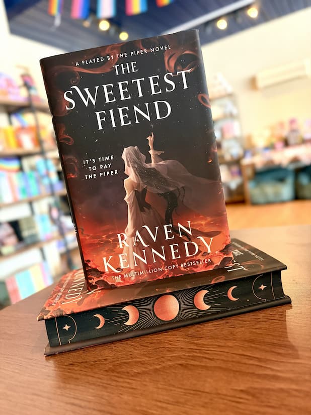 A small stack of the special edition of 'The Sweetest Fiend' by Raven Kennedy, showing a dark fantasy illustration of a hooded figure with a glowing staff against a fiery red and black background, with sprayed edges featuring moon phases.