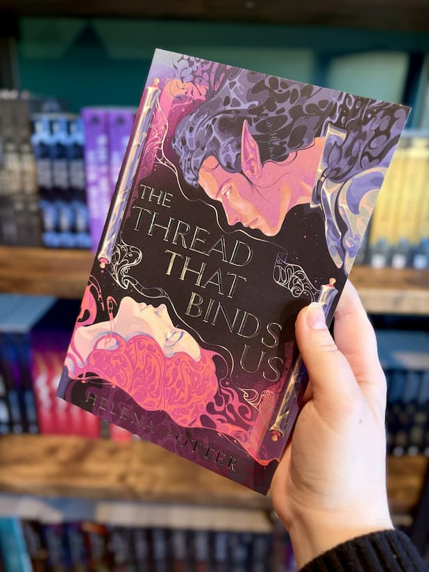 A hand holding a copy of 'The Thread That Binds Us' by Helena Potter, featuring an illustrated dark-haired male elf in profile and a pink-haired woman below, with ornate decorative elements against a black and purple background.