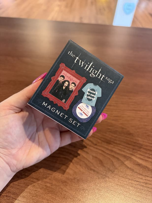 A hand with pink nail polish holds a square black box of 'The Twilight Saga' magnet set on a wooden table. The box features character artwork and a small t-shirt magnet illustration.