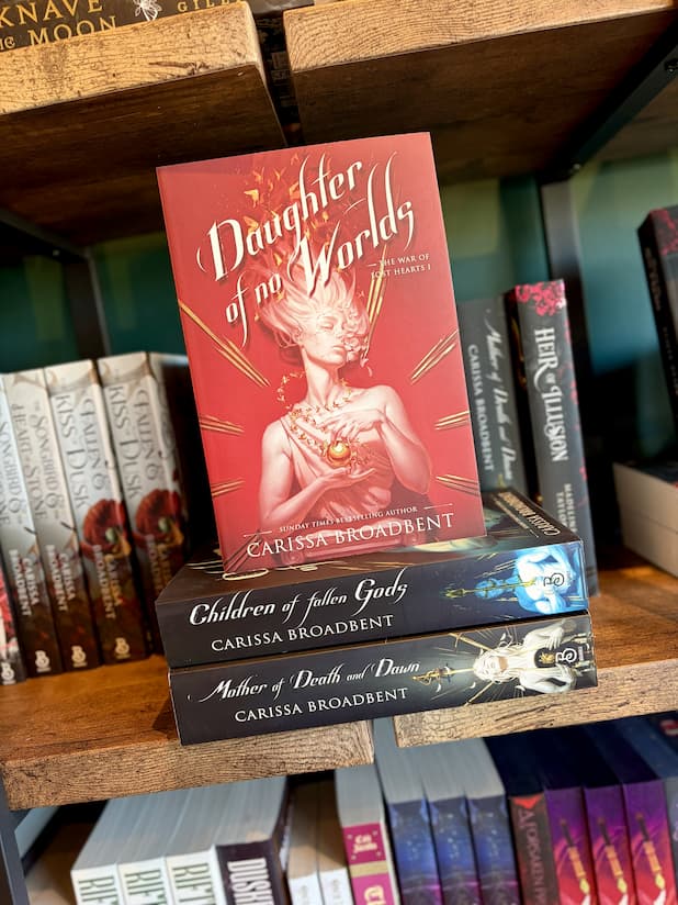 The book 'Daughter of No Worlds' by Carissa Broadbent is displayed upright on a wooden bookshelf, resting on top of two other books: 'Children of Fallen Gods' and 'Mother of Death and Dust'.