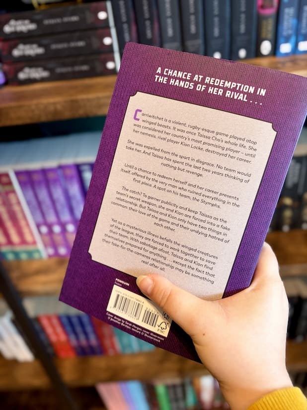 A hand holding the back cover of a book titled 'The Winged Game' by Sophie Kim, with a blurred bookshelf background. The book cover is purple with a silver and white winged emblem in the center.