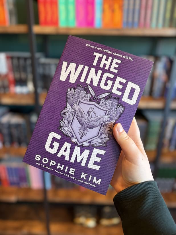 A hand holding a book titled 'The Winged Game' by Sophie Kim, with a blurred bookshelf background. The book cover is purple with a silver and white winged emblem in the center.