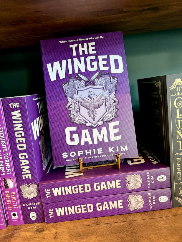 'The Winged Game' book by Sophie Kim displayed on a wooden shelf, with several copies of the book stacked around it. The book cover is purple with a silver and white winged emblem in the center.