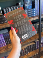 A hand with pink nail polish holds the back of a paperback copy of 'The Wolf and the Crown of Blood' by Elizabeth May, featuring a black background with red leaves along with the sides and white text.