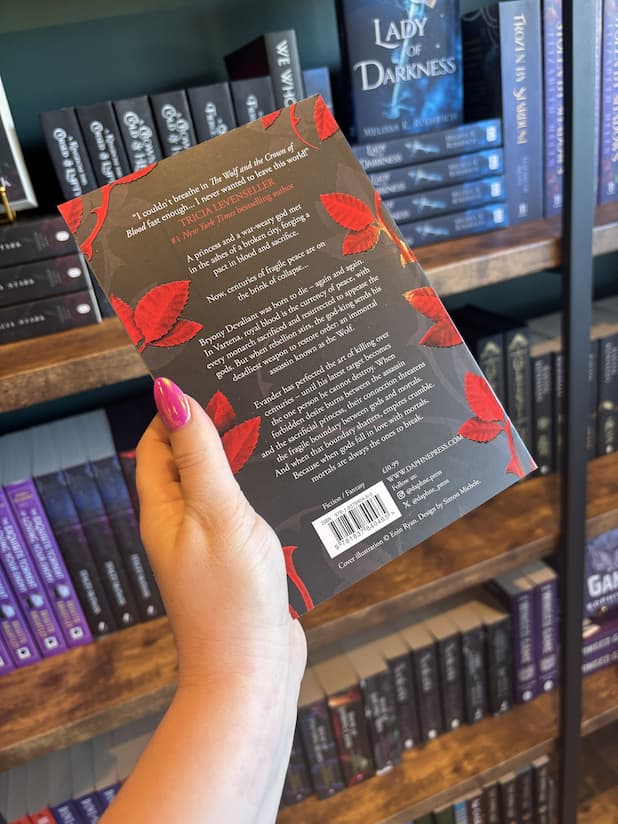 A hand with pink nail polish holds the back of a paperback copy of 'The Wolf and the Crown of Blood' by Elizabeth May, featuring a black background with red leaves along with the sides and white text.