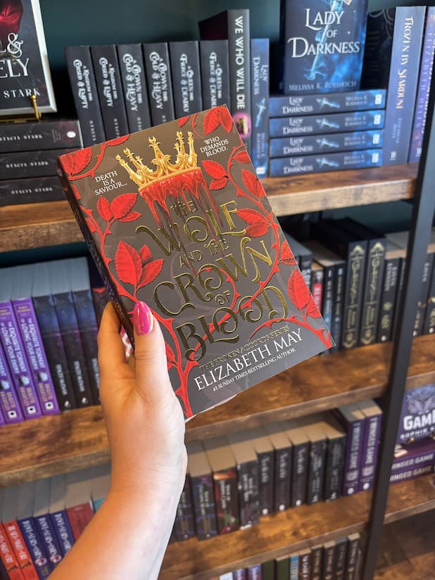A hand with pink nail polish holds a paperback copy of 'The Wolf and the Crown of Blood' by Elizabeth May, featuring a black background with red rose motifs, red blood dripping out of a gold crown, and large gold title text.