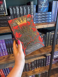 A hand with pink nail polish holds a paperback copy of 'The Wolf and the Crown of Blood' by Elizabeth May, featuring a black background with red rose motifs, red blood dripping out of a gold crown, and large gold title text.