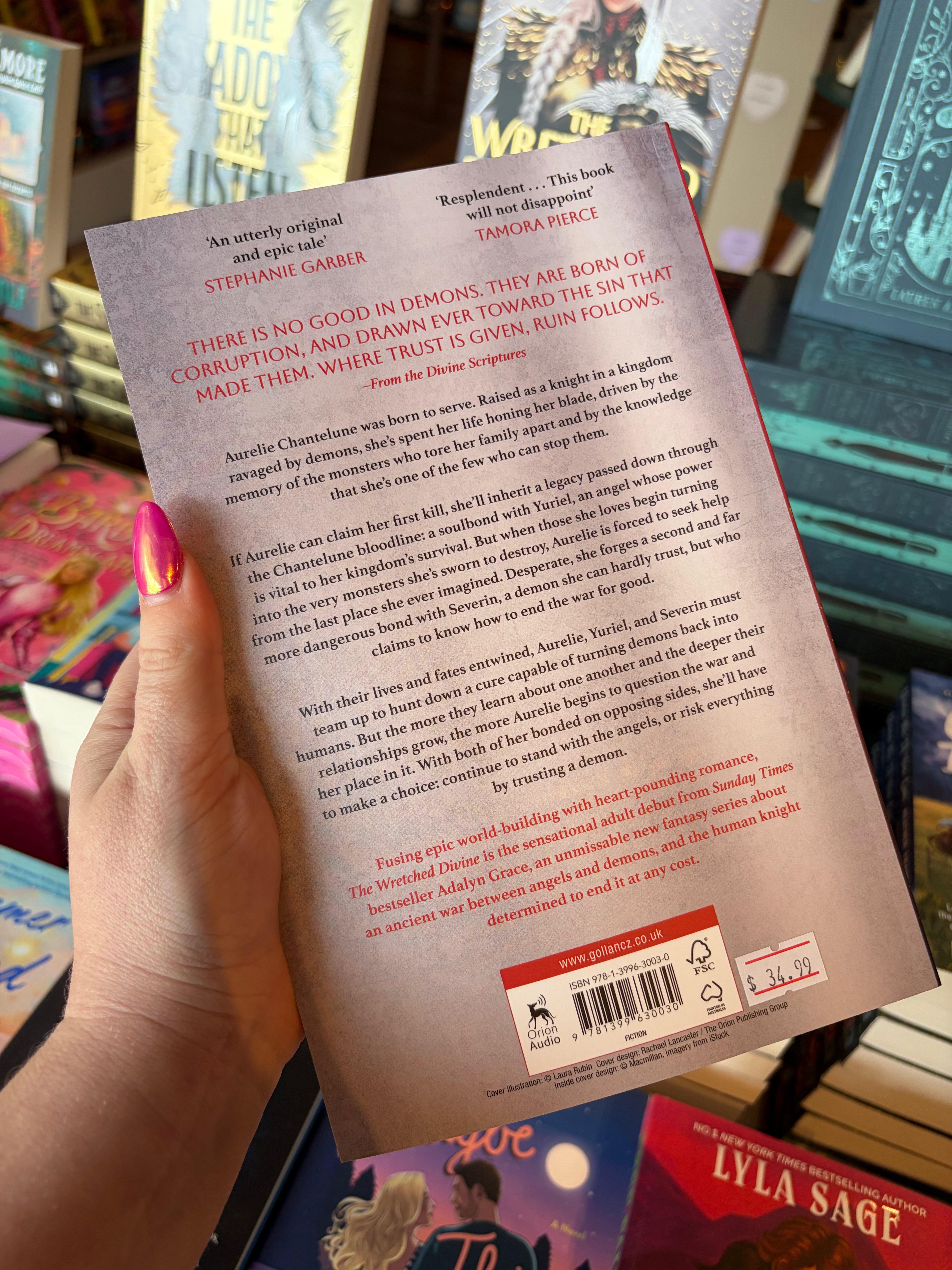 A hand holding the back cover of the book The Wretched Divine by Adalyn Grace, showing the title, reviews, and barcode against a colorful bookstore background.