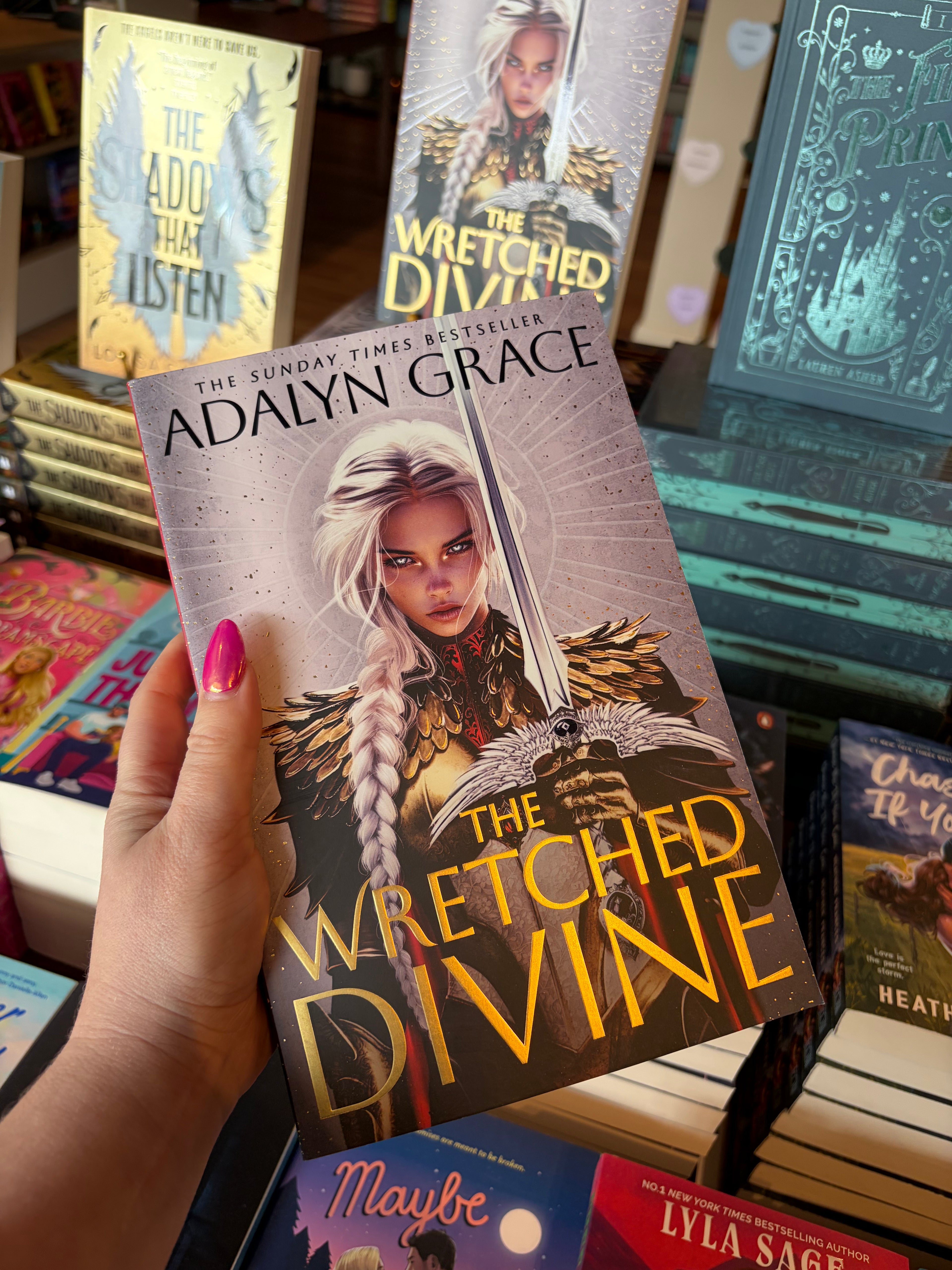 A hand with pink nail polish holds a copy of the book 'The Wretched Divine' by Adalyn Grace in a bookstore setting, surrounded by other book spines and covers.