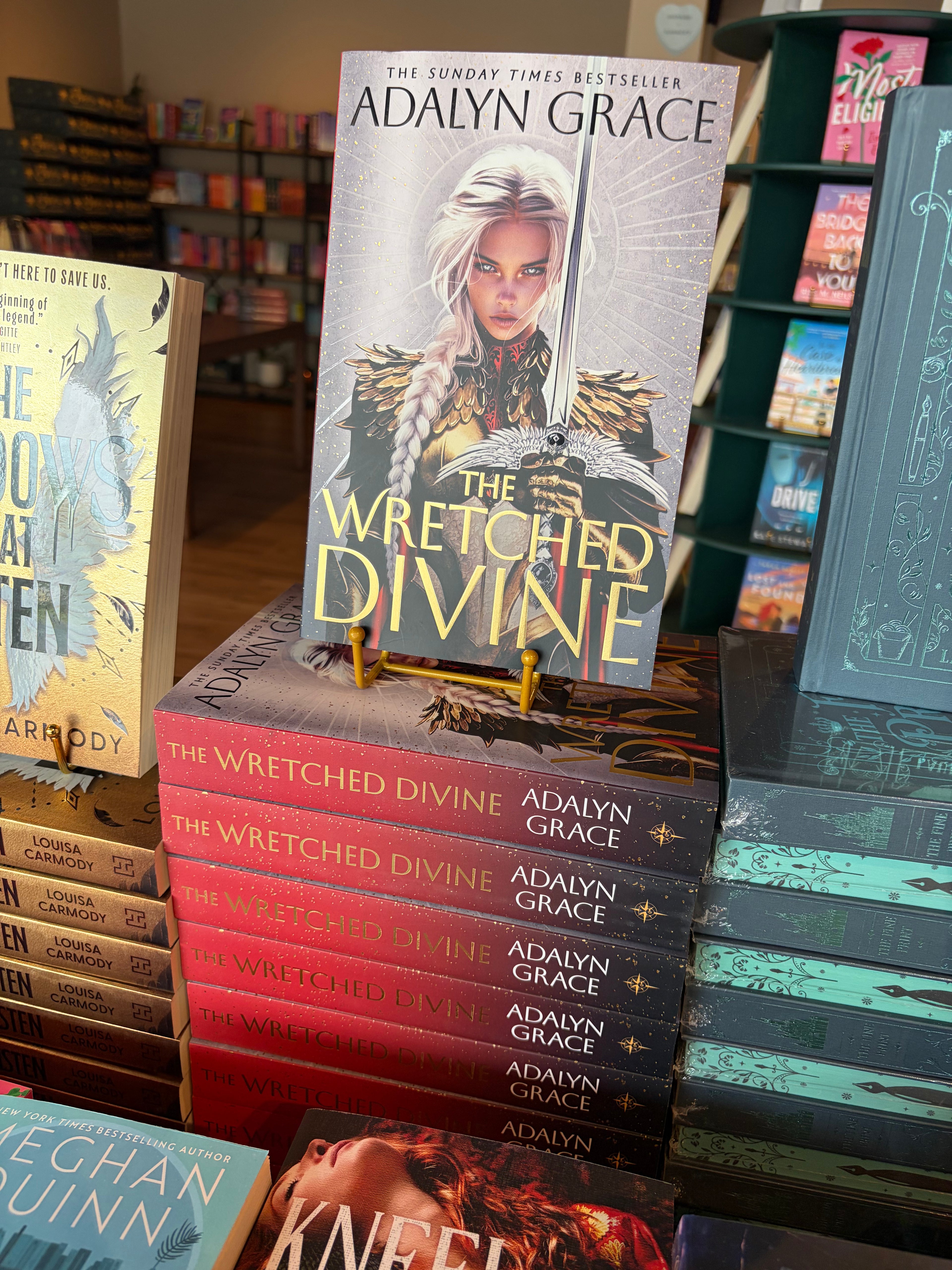 A stack of the book The Wretched Divine by Adalyn Grace in a bookstore with other books visible in the background. The cover features a female warrior with long white hair tied in a braid carrying a large sword. 