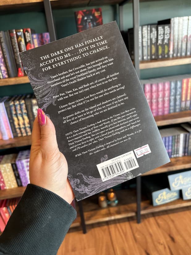 A hand with pink nail polish holding the back of a copy of 'Their Vicious Darling' by Nikki St. Crowe, featuring a textured black background and white text.