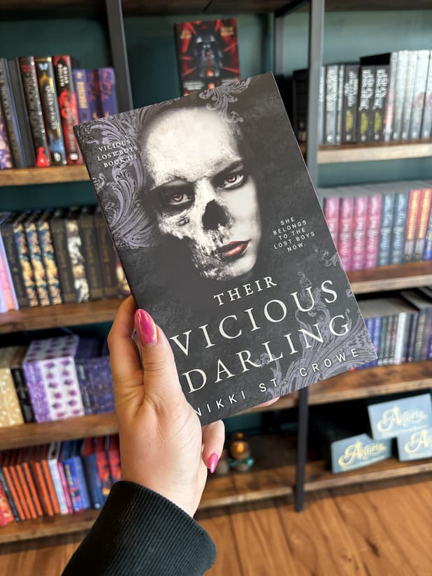 A hand with pink nail polish holding a copy of 'Their Vicious Darling' by Nikki St. Crowe, featuring a textured black background,a female face that is half a skull, and white text.