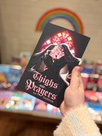 A hand holds a book titled 'Thighs and Prayers', Book One from the Tighs and Prayers series by Kat Blackthorne. The book cover is black with pink gothic-style lettering and an illustration of two women kissing under a stained-glass cathedral window.