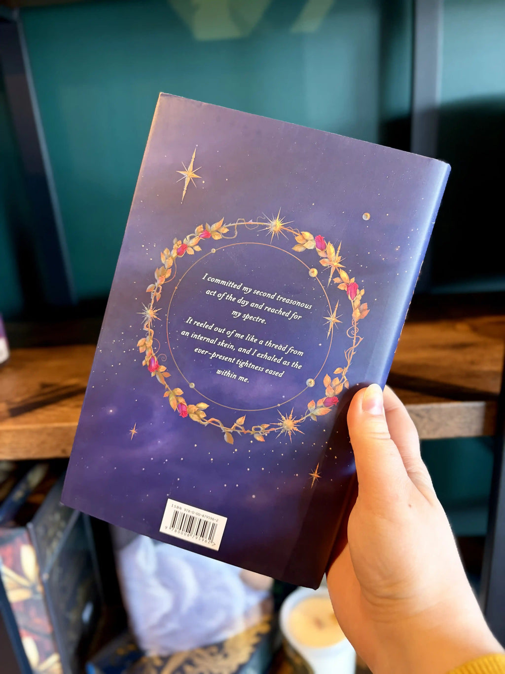 A hand holding the back of a paperback copy of the book 'Thorn Season' by Kiera Azar. The book cover features a dark purple background with a golden and red wreath, stars, and white text.